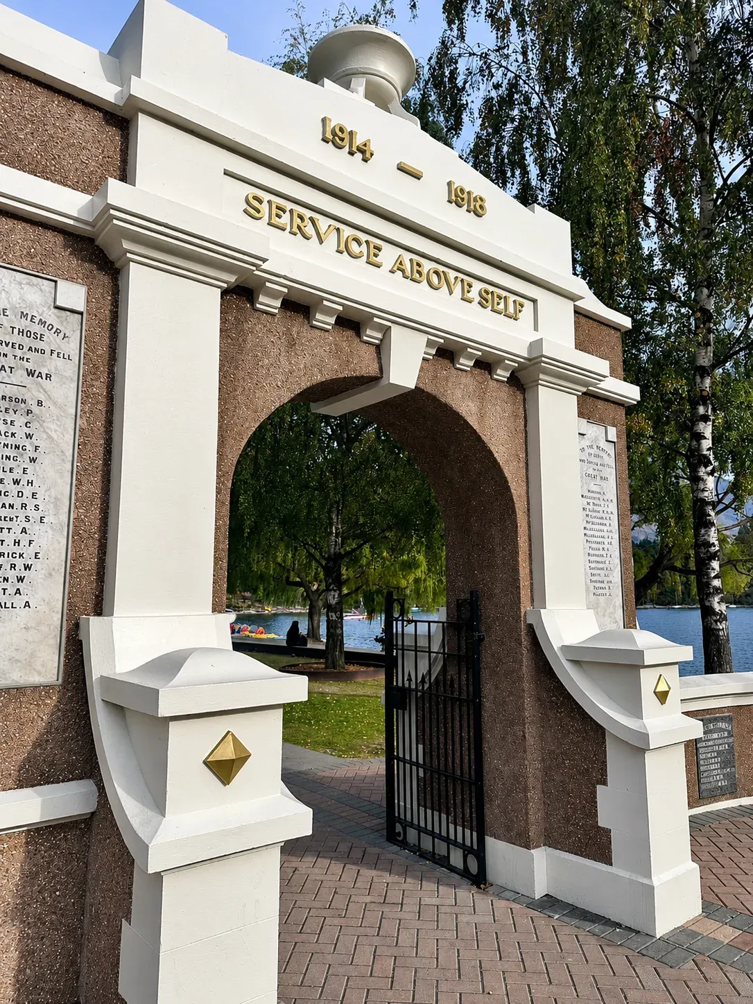 The Queenstown War Memorial archway, cream plaster trims and name panels either side of black steel gates, with trees beyond