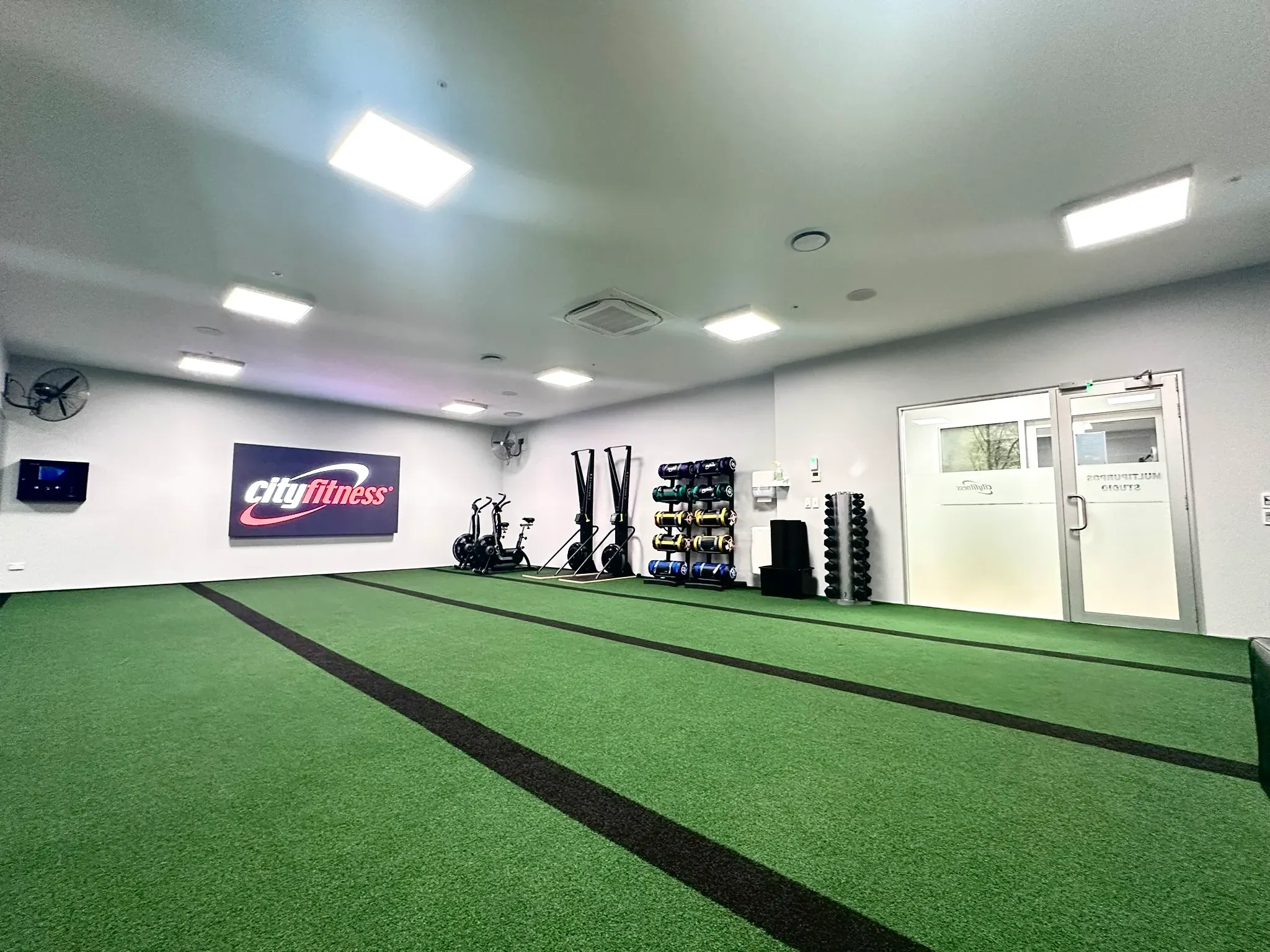 The gym floor at City Fitness Blenheim, a green turf sprint lane beside racked equipment under a pale painted ceiling