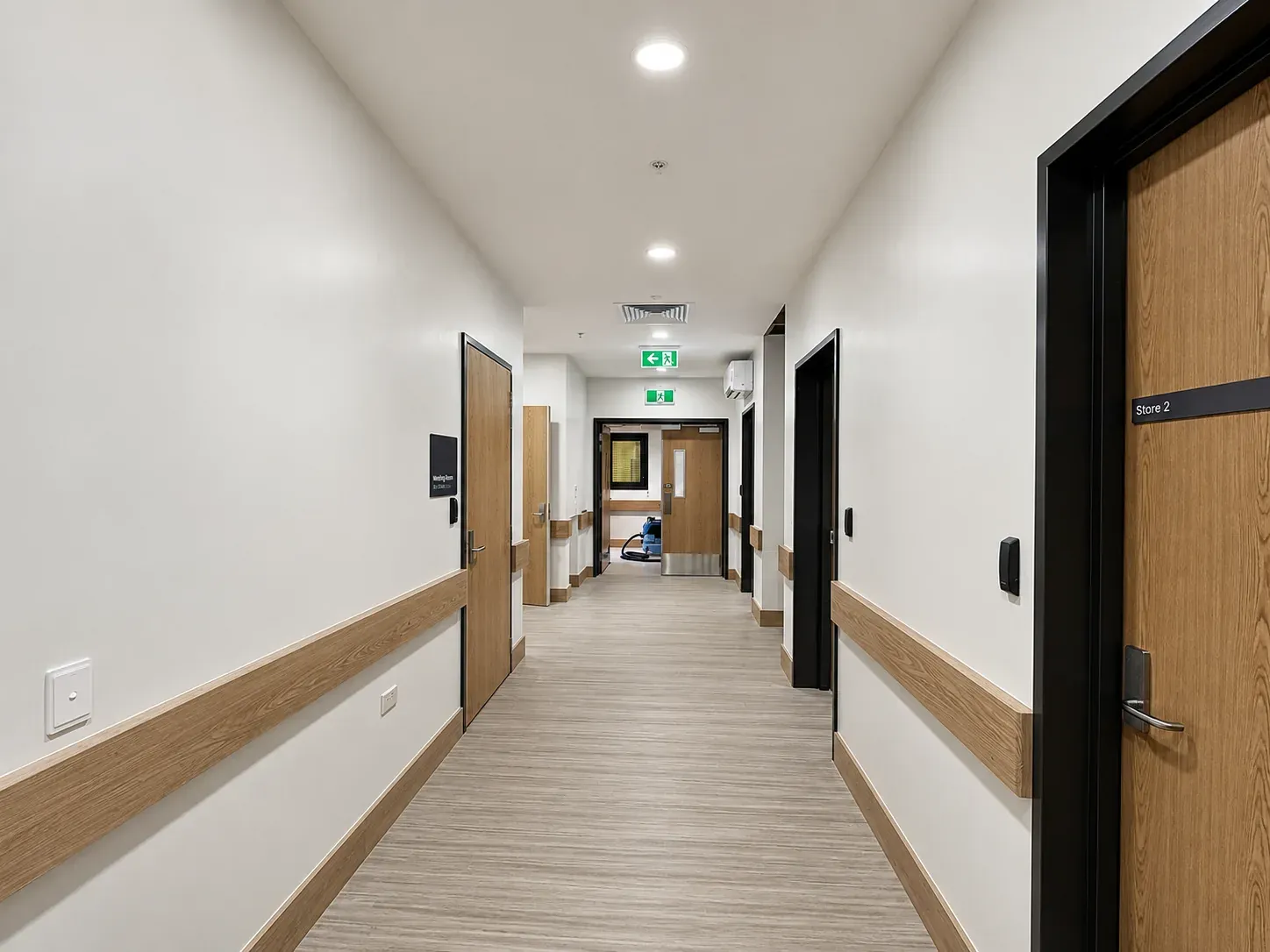 A corridor in the West Coast mental health facility, oak crash rails along pale walls with timber-veneer doors either side