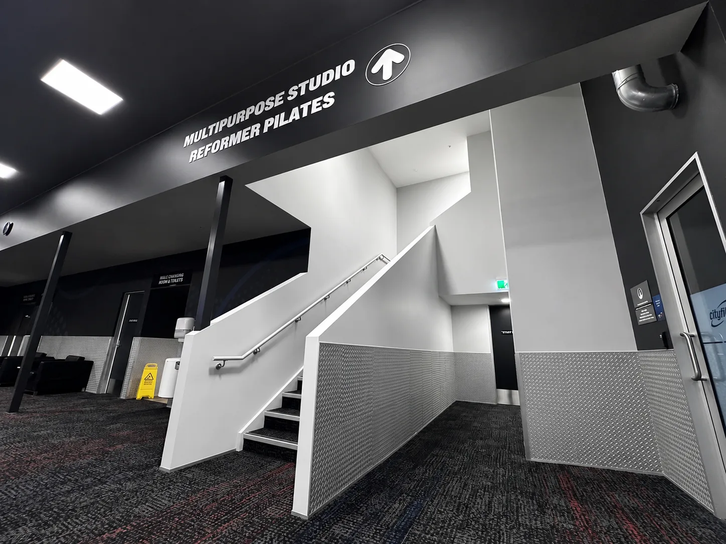 The stairwell up to the multipurpose and reformer pilates studios, white walls and handrail against dark ceiling and signage