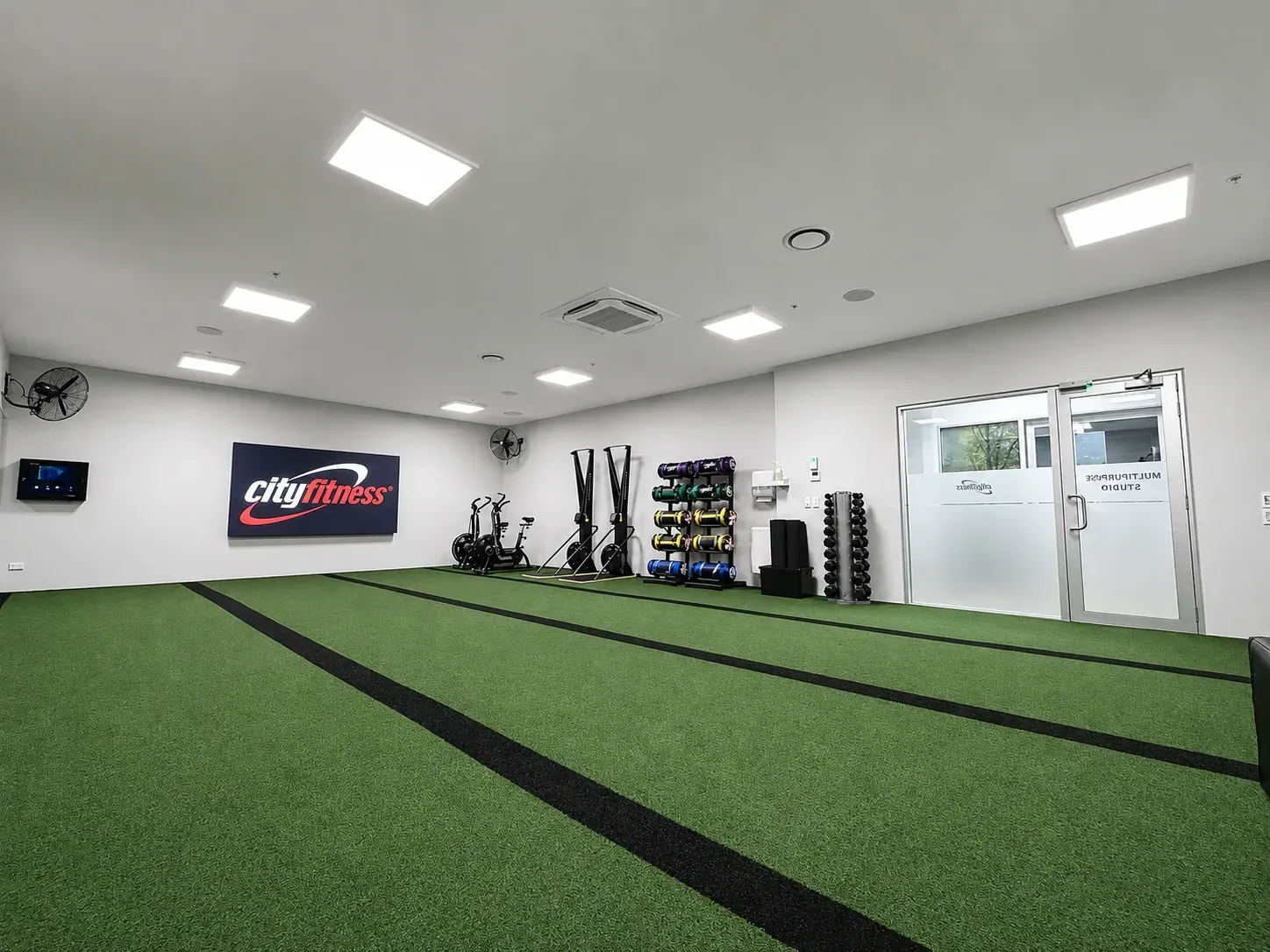 A wider view of the turf training floor, air bikes and dumbbell racks along a white wall beside the glazed studio doors