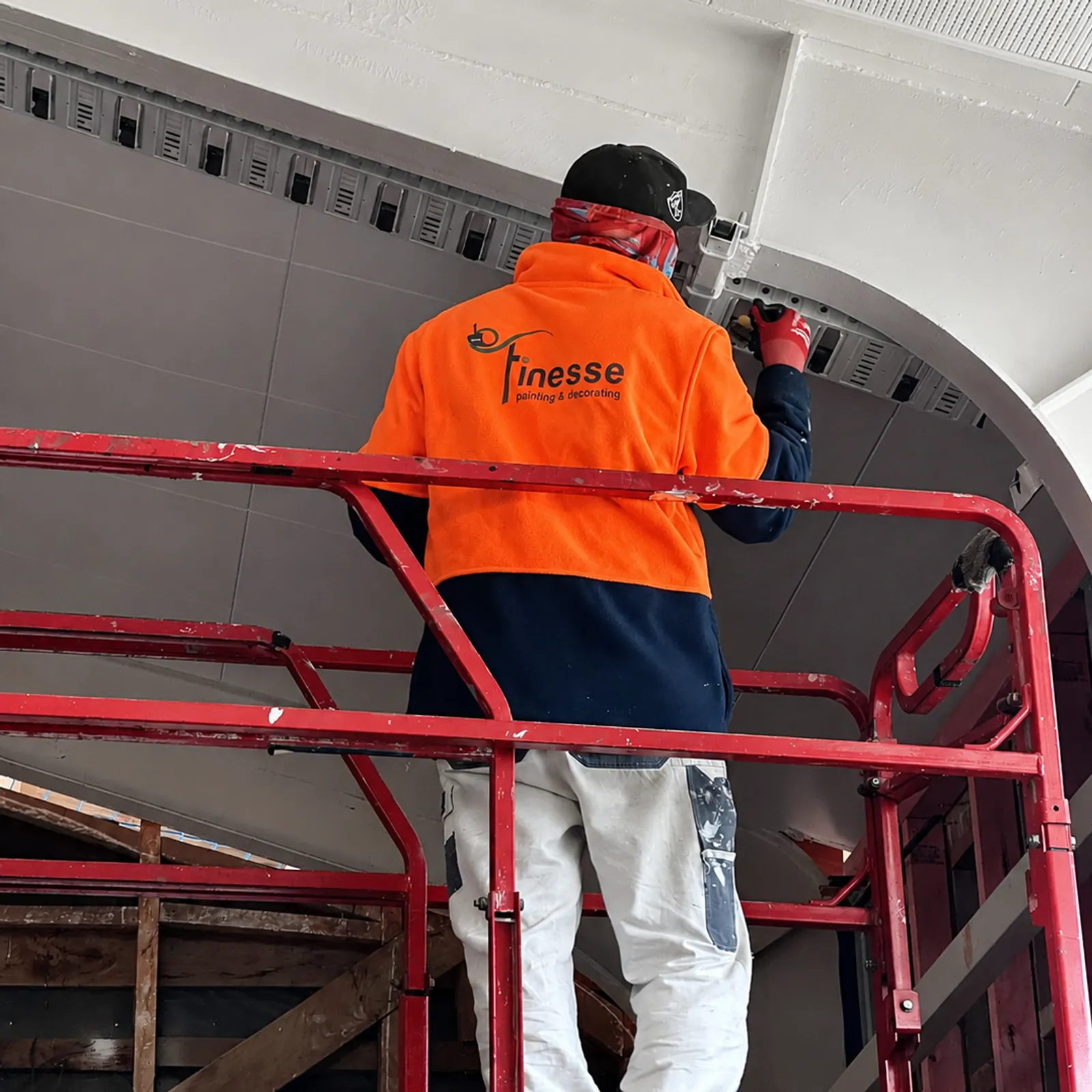 A Finesse painter in branded hi-vis working from a scissor lift, cutting in against the curved soffit of a commercial fit-out