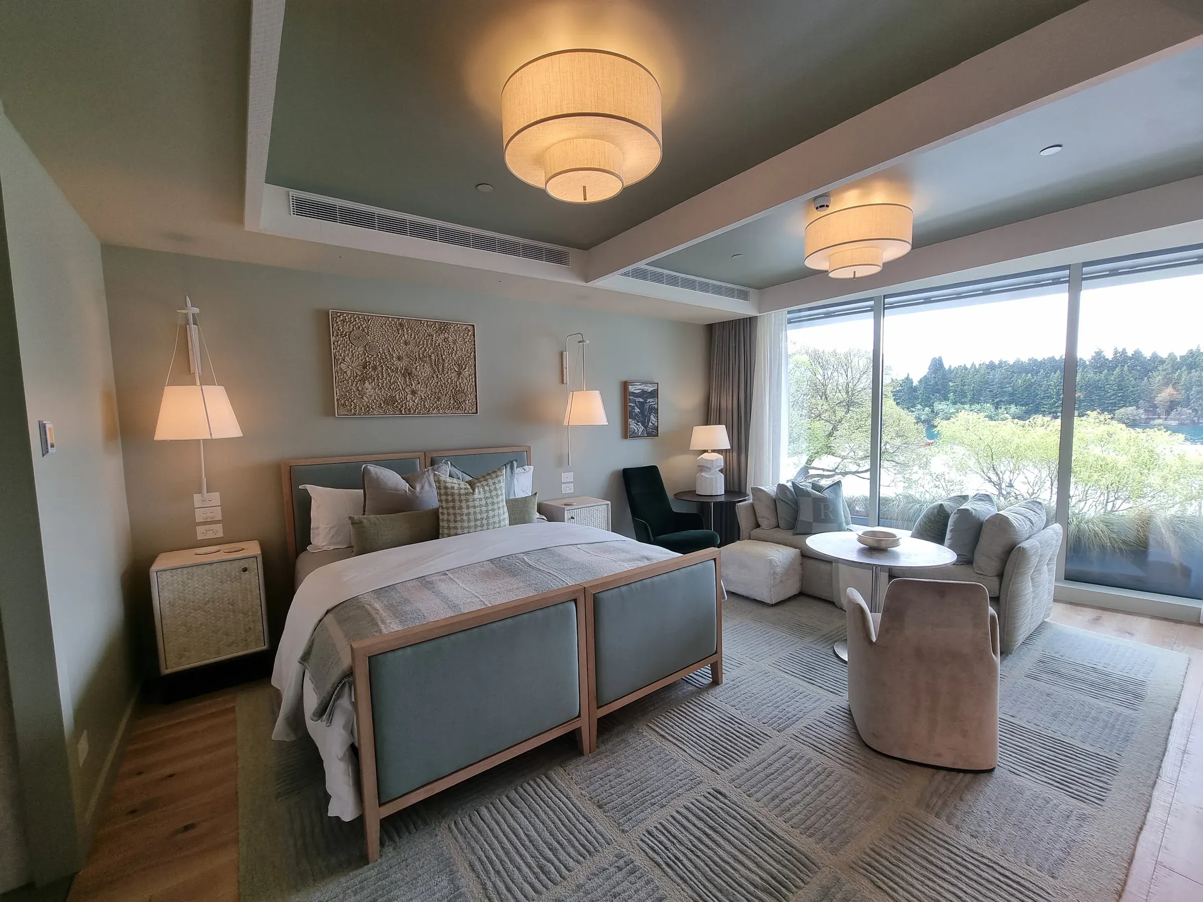 A Roki Collection bedroom suite, pale walls beneath a soft green-grey ceiling, with a sitting area facing the lake through full-height glazing