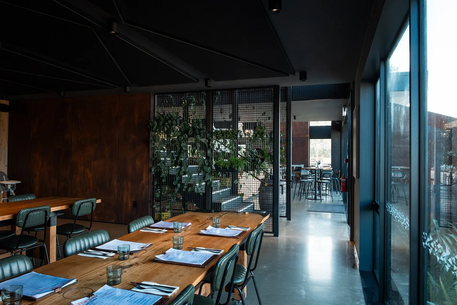 The Fancy Cow dining room, long timber tables and green leather chairs beside a rust-effect wall and a planted mesh screen