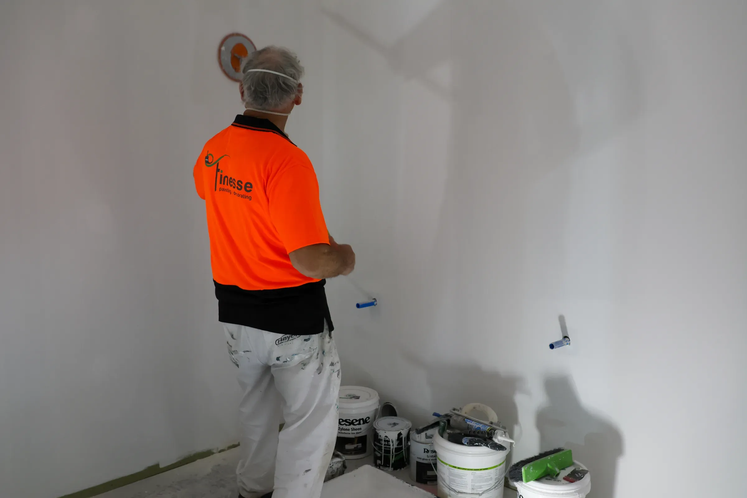 A Finesse painter in branded hi-vis rolling a freshly stopped interior wall, Resene tins and tools at his feet