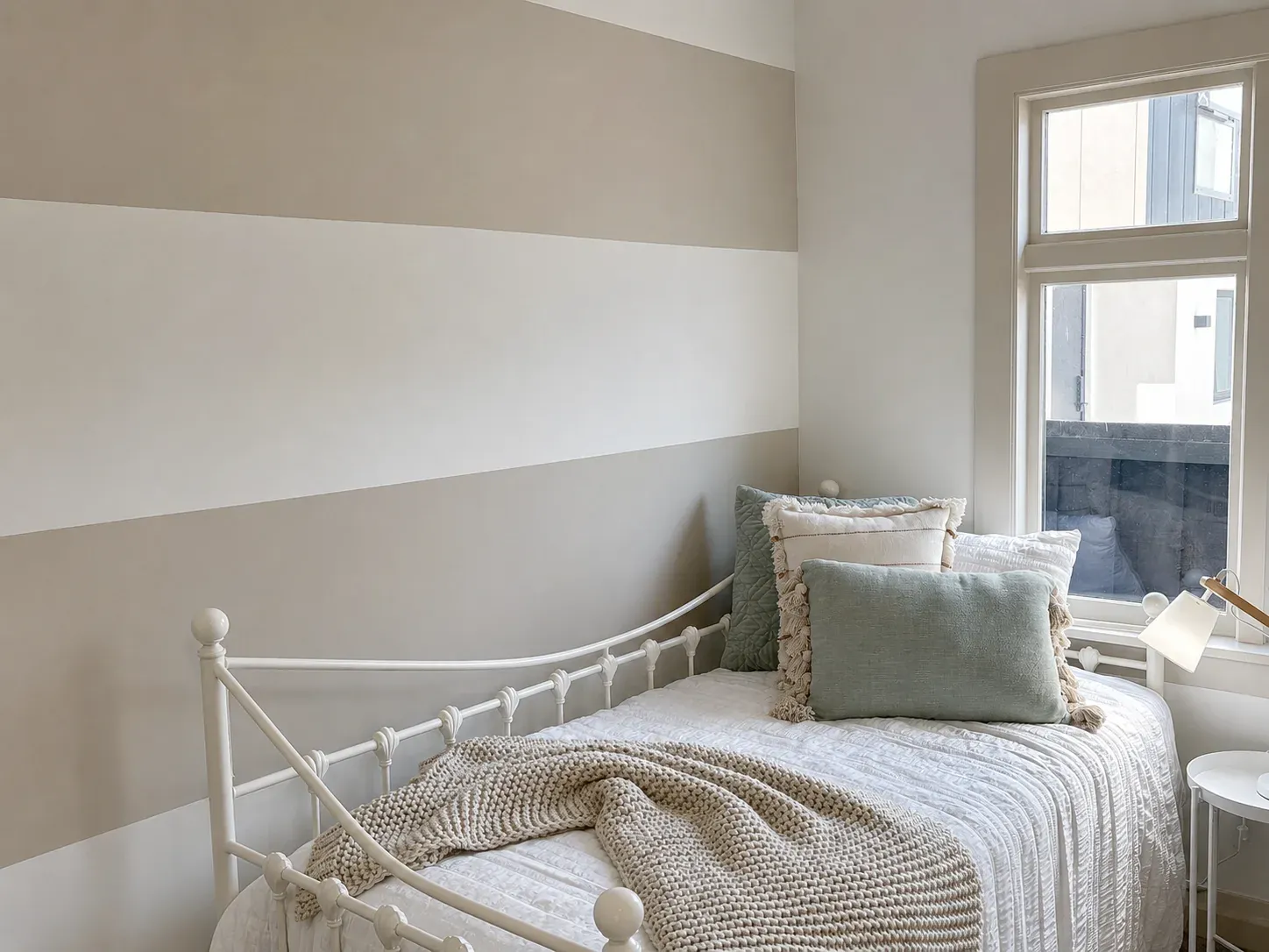 A residential bedroom with a soft two-tone striped feature wall, a white iron day bed made up in neutral linens