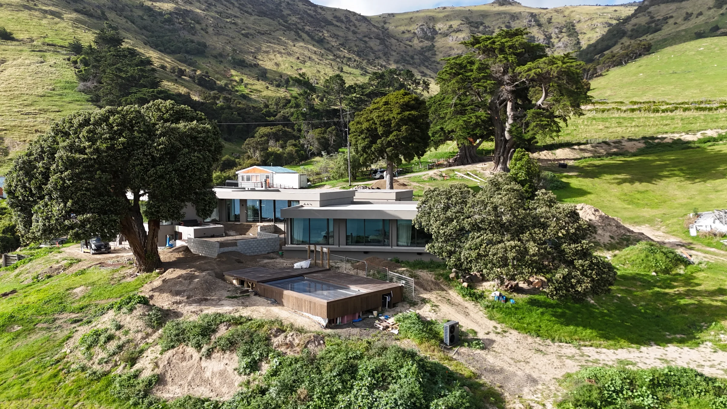 A low modern home on the Banks Peninsula coast, set among mature trees below steep green hills