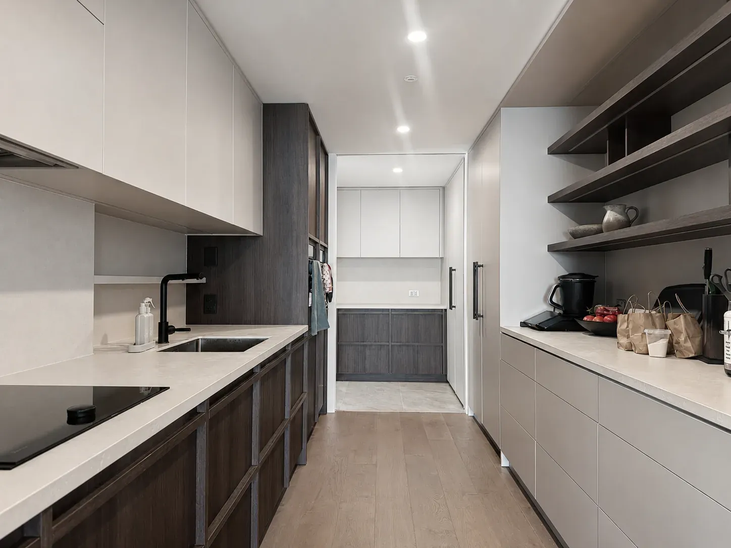 A galley kitchen at Little Akaloa, pale stone benchtops running between white and dark timber cabinetry, opening to a scullery beyond