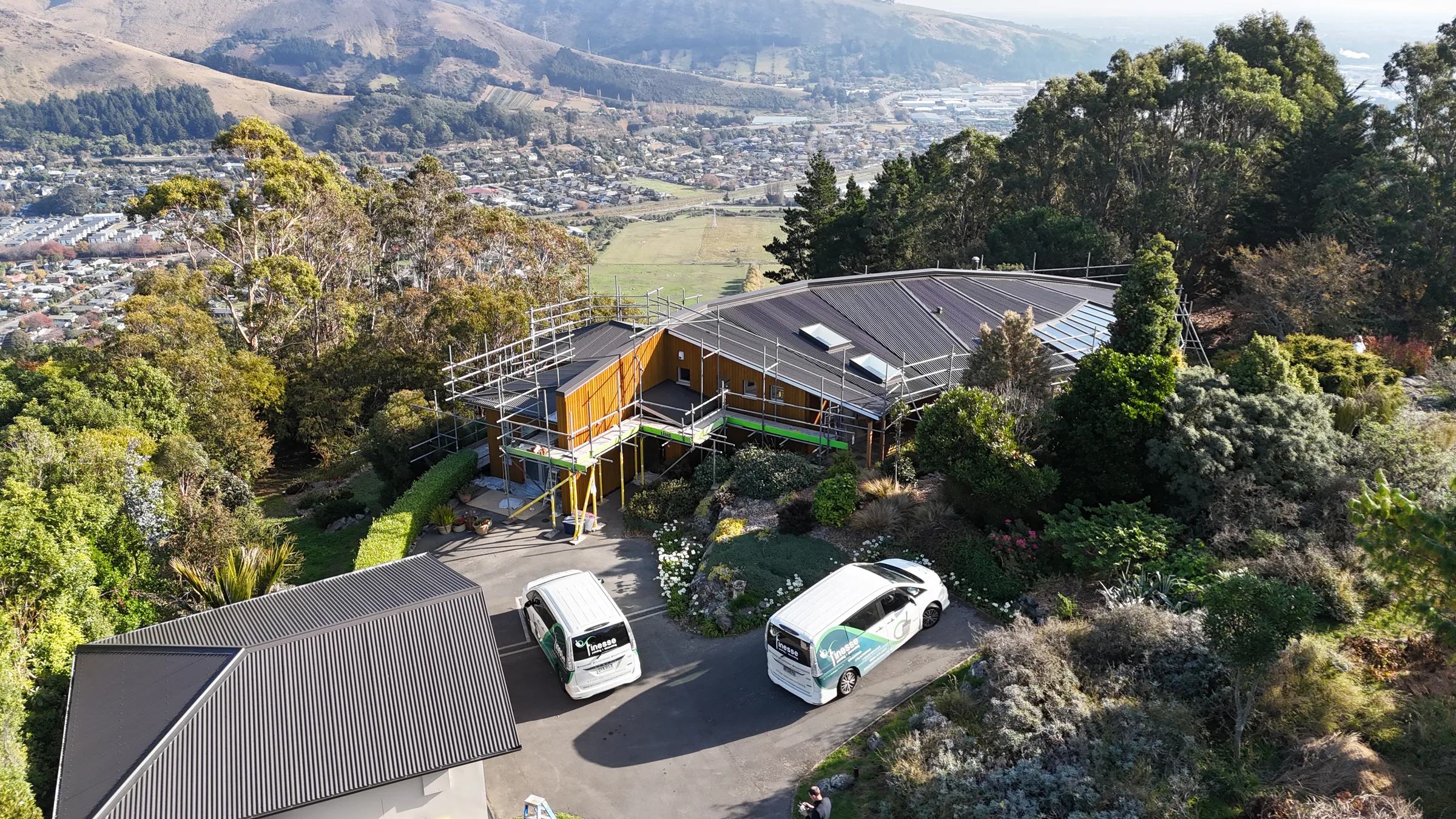 An aerial view of Rockview Terrace mid-repaint, a curved-roof home under scaffolding on a hillside above Christchurch, with two Finesse vans in the drive